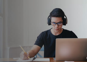 Man taking notes while using a laptop and wearing headphones